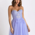 A woman poses in a strapless lavender gown with a fitted bodice featuring intricate lace detailing and a flowing tulle skirt by HADLEY PO926. The dress, model PO926, has a sweetheart neckline, and she is standing against a plain white background. Angels Formal Wear