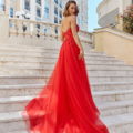 A woman in a vibrant red HADLEY PO926 gown poses elegantly on a set of white stone steps. The dress has a flowing train and a strapless bodice. She has a wavy bob haircut and is glancing back over her shoulder. The background features a multi-story building and balustrades. Angels Formal Wear