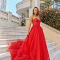 A woman in a flowing red gown stands on a staircase with ornate railings and the HADLEY PO926 building in the background. The dress features a strapless design and a long train. She is outdoors, surrounded by greenery and architectural features, creating an elegant scene that captures the essence of HADLEY PO926. Angels Formal Wear