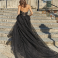 A woman with wavy brown hair, identified by the code PO926, is seen from behind walking up stone steps in an outdoor setting. She is wearing a strapless, glittery black gown with a long, flowing train that sweeps the ground. A multi-level building by HADLEY PO926 is visible in the background. Angels Formal Wear