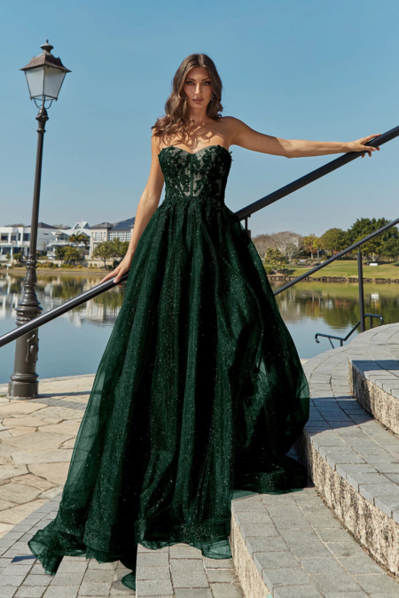 A woman in a flowing dark green gown stands on outdoor steps by a scenic lakeside, the strapless dress featuring a fitted bodice and voluminous skirt. She has long, wavy hair and gazes into the distance under a clear blue sky. The scene is graced by a vintage streetlamp, trees, and the timeless elegance of HADLEY PO926. Angels Formal Wear