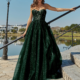 A woman in a flowing dark green gown stands on outdoor steps by a scenic lakeside, the strapless dress featuring a fitted bodice and voluminous skirt. She has long, wavy hair and gazes into the distance under a clear blue sky. The scene is graced by a vintage streetlamp, trees, and the timeless elegance of HADLEY PO926. Angels Formal Wear