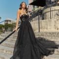 A woman in an elegant, sparkling black strapless gown stands on an outdoor staircase. She has wavy brown hair and is posing with her hand on the railing, with buildings and railings in the background under a clear blue sky. The HADLEY PO926 dress adds to her timeless allure. Angels Formal Wear