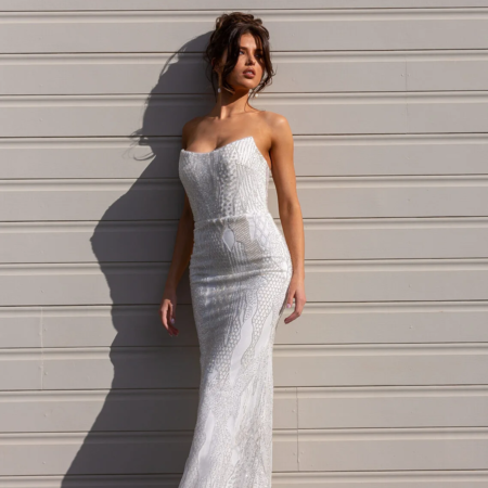 Woman wearing a strapless, form-fitting white gown, reminiscent of the NORA PO974 design, stands against a light-colored, ribbed wall. The floor-length dress has a textured pattern. She gazes to the side, her shadow cast prominently against the wall. The ground beneath is gravel. Angels Formal Wear