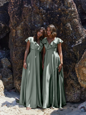 Two women in long, silky green dresses stand barefoot against a backdrop of large, rugged rocks on a sandy beach. Both have shoulder-length brown hair and are gazing forward with relaxed and serene expressions. The scene is as tranquil as the melody of PETAL TO882 playing softly in the background. Angels Formal Wear