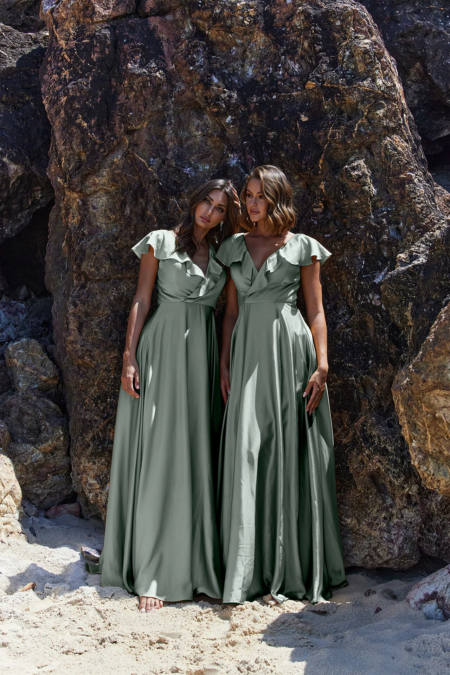 Two women in long, silky green dresses stand barefoot against a backdrop of large, rugged rocks on a sandy beach. Both have shoulder-length brown hair and are gazing forward with relaxed and serene expressions. The scene is as tranquil as the melody of PETAL TO882 playing softly in the background. Angels Formal Wear