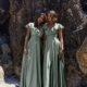 Two women in long, silky green dresses stand barefoot against a backdrop of large, rugged rocks on a sandy beach. Both have shoulder-length brown hair and are gazing forward with relaxed and serene expressions. The scene is as tranquil as the melody of PETAL TO882 playing softly in the background. Angels Formal Wear