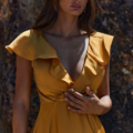 A woman with long, dark hair stands against a textured, rocky background. She is wearing a yellow PETAL TO882 with ruffled cap sleeves and a deep V-neckline that resembles the soft curve of a petal. Her expression is calm and composed, and her left hand rests lightly on her dress. Angels Formal Wear