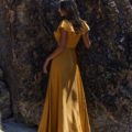 A woman stands on a sandy beach with her back to the camera. She is wearing a long, flowing, mustard-yellow dress with ruffled sleeves. She is leaning against a large, rocky formation as her long hair cascades down her back, resembling petals in motion captured by the PETAL TO882. Angels Formal Wear