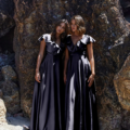 Two women stand barefoot on sand in front of large rocks, wearing matching black satin dresses with ruffled sleeves. Their shoulder-length hair frames their faces as they stand close to each other, exuding a serene and confident presence. One of the dresses appears to be a PETAL TO882 design. Angels Formal Wear