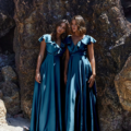 Two women stand side by side on a beach, wearing matching, elegant blue PETAL TO882 satin dresses with ruffled sleeves. They pose in front of large, rocky formations, with sand at their feet and sunlight illuminating their outfits. Angels Formal Wear