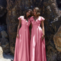 Two women in matching pink PETAL TO882 floor-length dresses stand closely together on a sandy beach, with large rocky formations behind them. Both have dark hair and are gazing directly at the camera, exuding a serene and confident demeanor. Angels Formal Wear