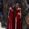 Two women in matching flowing PETAL TO882 dresses stand side by side against a large rocky backdrop on a beach. Both are barefoot, and their hair is styled similarly. The natural sunlight highlights the sheen of their elegant gowns, reminiscent of rose petals, and the rough texture of the rocks. Angels Formal Wear