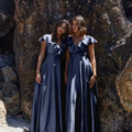 Two women in long, dark blue PETAL TO882 satin dresses with petal-like ruffled sleeves stand close together on a sandy beach, leaning against large, textured rocks. They exhibit a serene, relaxed demeanor. Angels Formal Wear