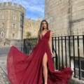 A woman in a flowing deep red dress poses confidently on a cobblestone street. Behind her, there is a historic stone building with a tower and arched windows, bordered by a black iron fence. The clear blue sky frames the scene perfectly, capturing the essence of PHOEBE JP124 elegance. Angels Formal Wear