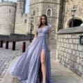 A woman in a flowing, light blue gown stands on a stone pathway next to the historic PHOEBE JP124 castle with tall, cylindrical towers. She has long hair, wears white sandals, and poses with one leg slightly extended from a thigh-high slit in her dress. Angels Formal Wear