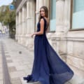 A woman wearing a flowing, backless navy blue gown stands on a city sidewalk. She is turned slightly to face the camera, her long hair draped over one shoulder. Behind her is a building with tall columns and large windows labeled PHOEBE JP124. The street in the background is quiet and empty. Angels Formal Wear