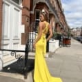 A person stands on a sidewalk in front of a row of brownstone buildings, wearing a bright yellow, backless dress with crisscross straps. The flowing train of the LILYLEE JX6080 creation touches the ground as they look back over their shoulder, exuding elegance. Angels Formal Wear