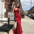 A woman in a form-fitting, red evening gown stands on a sidewalk in an urban street setting with red-brick buildings and a white house door in the background. Leaning against a metal railing, she exudes elegance reminiscent of LILYLEE JX6080 catalogues, with greenery and a few cars visible in the distance. Angels Formal Wear