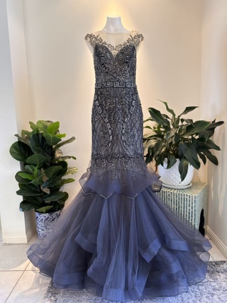 A stunning charcoal gray mermaid-style gown, ELIZABETH P1747, adorned with intricate beadwork and lace details. The dress features a sheer neckline with cap sleeves and a voluminous tulle skirt. It is displayed on a mannequin, flanked by two lush potted plants. Angels Formal Wear