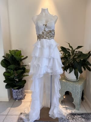 A white strapless GINA is displayed on a mannequin in a well-lit room. The dress features a textured bodice with gold embellishments and a layered, ruffled skirt with a high-low hemline. There are potted plants on either side of the mannequin. Angels Formal Wear