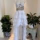 A white strapless GINA is displayed on a mannequin in a well-lit room. The dress features a textured bodice with gold embellishments and a layered, ruffled skirt with a high-low hemline. There are potted plants on either side of the mannequin. Angels Formal Wear