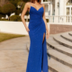 A woman stands outdoors in front of a white building with stairs, wearing a floor-length, royal blue evening gown with a thigh-high slit. The AUTO-DRAFT dress is sleeveless with thin straps and features ruched, sparkling fabric. She has long, wavy hair and is wearing sandals. Angels Formal Wear