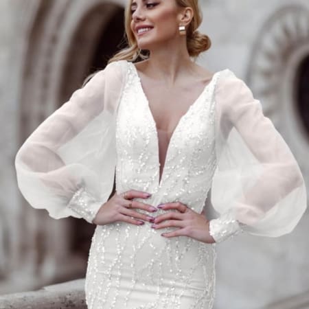 A smiling woman in a white bridal gown, NINA TK209W, stands outdoors. The dress features long, puff sleeves and intricate beading. Her hair is styled in a side-swept updo, and she is looking away from the camera. An architectural background with arched detailing is visible. Angels Formal Wear