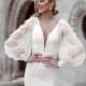 A smiling woman in a white bridal gown, NINA TK209W, stands outdoors. The dress features long, puff sleeves and intricate beading. Her hair is styled in a side-swept updo, and she is looking away from the camera. An architectural background with arched detailing is visible. Angels Formal Wear