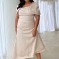 A woman wearing a blush pink ARITA TL DRESS with an asymmetrical neckline and cap sleeves. She has long, dark hair, is standing in front of sheer white curtains and a white wall, and is wearing strappy silver heels. A decorative floral arrangement is visible overhead. Angels Formal Wear