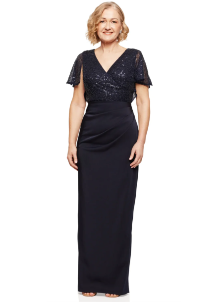 A woman with short blonde hair is standing and smiling, wearing the elegant JOYCE DRESS - TL1063PS in navy blue—a floor-length gown with sequined short sleeves and a V-neck. The background is plain white. Angels Formal Wear