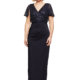 A woman with short blonde hair is standing and smiling, wearing the elegant JOYCE DRESS - TL1063PS in navy blue—a floor-length gown with sequined short sleeves and a V-neck. The background is plain white. Angels Formal Wear