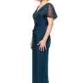 A woman with short, wavy blonde hair is standing and smiling in a stunning JOYCE DRESS - TL1063PS. She wears a floor-length navy blue gown featuring a sequin-embellished bodice and sheer short sleeves. She is facing slightly to the right, with her left side visible in the image. Angels Formal Wear