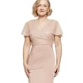 A woman with short, curly blonde hair wearing the stunning JOYCE DRESS - TL1063PS, a blush pink, short-sleeved gown, smiles and stands against a plain white background. The gown has a V-neck and is fitted at the waist with subtle shimmering details on the top. Angels Formal Wear