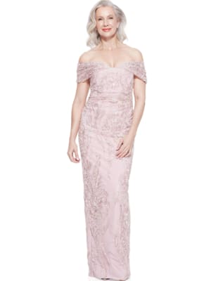 A smiling woman with gray hair is wearing an elegant, off-the-shoulder, light pink ALIYA DRESS - TL1114 with intricate beadwork. She is standing against a plain white background. Angels Formal Wear