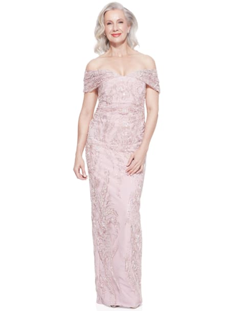 A smiling woman with gray hair is wearing an elegant, off-the-shoulder, light pink ALIYA DRESS - TL1114 with intricate beadwork. She is standing against a plain white background. Angels Formal Wear