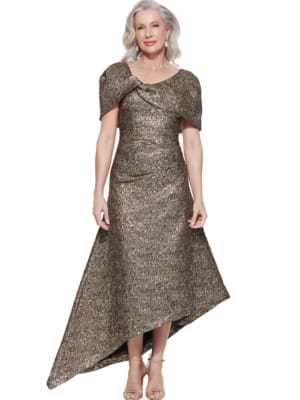 A woman with long, wavy gray hair is wearing a shimmering ARITA TL DRESS with an asymmetrical gold design and short sleeves. The dress features a unique shoulder detail and an uneven hemline. She stands on a white background, smiling at the camera. Angels Formal Wear