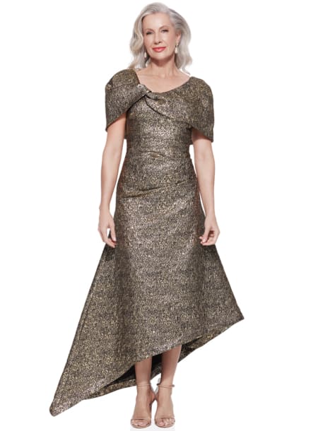 A woman with long, wavy gray hair is wearing a shimmering ARITA TL DRESS with an asymmetrical gold design and short sleeves. The dress features a unique shoulder detail and an uneven hemline. She stands on a white background, smiling at the camera. Angels Formal Wear