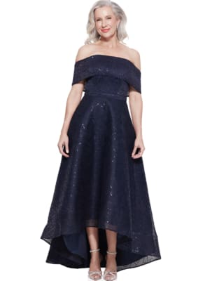 An elderly woman with shoulder-length grey hair is wearing an off-the-shoulder, navy blue ROSITA DRESS - TL1143 with intricate detailing. She has a slight smile and is standing against a plain white background, showing her silver strappy sandals. Angels Formal Wear