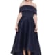 An elderly woman with shoulder-length grey hair is wearing an off-the-shoulder, navy blue ROSITA DRESS - TL1143 with intricate detailing. She has a slight smile and is standing against a plain white background, showing her silver strappy sandals. Angels Formal Wear