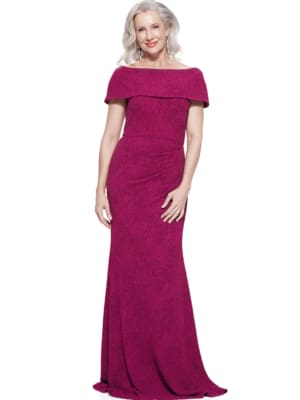 A woman named Shari with shoulder-length, wavy white hair stands posed, wearing a floor-length, off-the-shoulder deep magenta Shari Gown with a textured pattern. She is smiling, with her hands gently resting by her sides. Angels Formal Wear