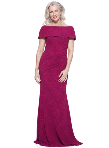 A woman named Shari with shoulder-length, wavy white hair stands posed, wearing a floor-length, off-the-shoulder deep magenta Shari Gown with a textured pattern. She is smiling, with her hands gently resting by her sides. Angels Formal Wear