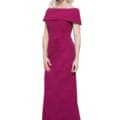 Shari, with her gray hair elegantly framing her face, wears a floor-length, off-the-shoulder fuchsia Shari Gown as she poses against a plain white background. She stands confidently with a gentle smile and her hands relaxed at her sides. Angels Formal Wear