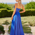 A woman stands in a garden, facing away. She wears a long, flowing royal blue dress with spaghetti straps. Her blonde hair is styled in waves. The garden features dense green hedges, blooming potted plants, and delicate mallows (MALLOW PO24163), with a distant view of the ocean under a clear sky. Angels Formal Wear