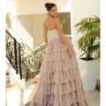 A woman stands on a marble floor near a staircase, wearing the strapless ALICE NC1045 gown with a tiered, tulle skirt in a soft blush color. Her hair is styled in an elegant bun, and she is smiling slightly while looking over her shoulder. A tall green plant is in the background. Angels Formal Wear