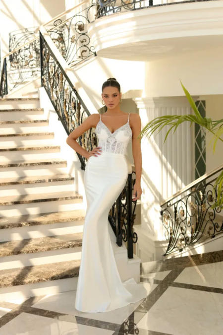 A woman in an elegant white gown stands on a staircase with ornate wrought iron railings. Sunlight filters through the space, casting shadows on the marble steps. She gazes directly at the camera, exuding confidence and grace, with an ALITZA NC1018 clutch in her hand. A palm frond is visible to the right.


 Angels Formal Wear