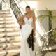 A woman in an elegant white gown stands on a staircase with ornate wrought iron railings. Sunlight filters through the space, casting shadows on the marble steps. She gazes directly at the camera, exuding confidence and grace, with an ALITZA NC1018 clutch in her hand. A palm frond is visible to the right.


 Angels Formal Wear