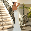 A woman stands on a marble staircase with intricate black railings, elegantly dressed in an ALITZA NC1018 gown featuring a form-fitting design and a delicate lace bodice. She gazes over her shoulder, her hair neatly styled up, while sunlight streams through a large window, casting soft shadows. Angels Formal Wear