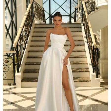 A woman in a strapless, white ANNA NC1076 (Copy) gown poses confidently on a grand staircase with intricate iron railings and a geometric glass backdrop. The gown features a thigh-high slit, and she pairs it with high heels, showcasing an elegant and sophisticated look. Angels Formal Wear