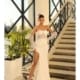 A woman wearing the ASHLEY NC1036, a white strapless gown with a high slit and sheer gloves, poses by a staircase with intricate railings and large windows in the background. Her stance, one hand resting on the railing and the other on her hip, exudes ASHLEY's signature elegance and confidence. Angels Formal Wear